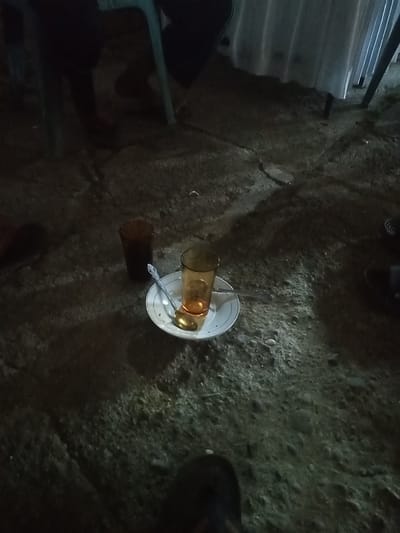 An overhead, low-light shot captures a ground-level view of a paved area in Ulee Breuk, Indonesia, likely an outdoor seating area at night. In the center, a white plate holds two glasses and a spoon. One glass, partially filled with a dark liquid, stands to the left. To the right, another glass, clearer and containing a lighter liquid, is also present. A silver spoon rests on the plate, positioned as if used for stirring. The ground is rough and uneven, with visible pebbles and dirt. Shadows cast by unseen objects and lighting create a dramatic atmosphere, obscuring much of the surrounding environment and any individuals present. The overall scene suggests a casual gathering or break in a dimly lit outdoor setting.