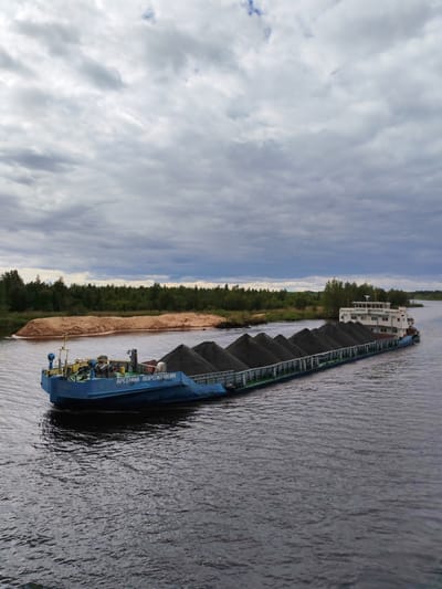 The image shows a blue barge filled with dark gray cargo, possibly coal or gravel, navigating a wide, calm river. The barge has the name "АРСЕНИЙ ВОРОЖЕЙКИН" written on its side in Cyrillic script. Behind the barge, a sandy embankment is visible on the left, topped with trees. A dense green forest lines the far bank of the river. The sky is overcast with dramatic gray clouds. The scene appears to be located in or near Ozernoe Ust'e, Russia, given the context of the provided location. The water is dark and reflects the cloudy sky. There are no people clearly visible in the image. The overall impression is one of industrial activity within a natural landscape.