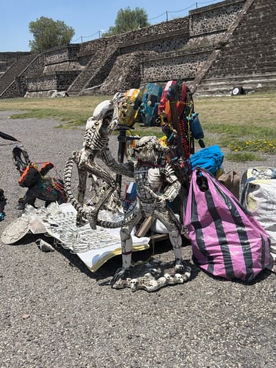The image depicts a collection of items set up on a paved surface, potentially for sale or display. Dominating the scene are two skeletal figures, one standing and one seated, both appearing to be crafted from bone or bone-like material. The seated figure is adorned with a variety of beads and trinkets. Behind the figures are several bags, including one with pink and black stripes. To the left, a stone pyramid structure is visible. The location is San Francisco Mazapa, Mexico.