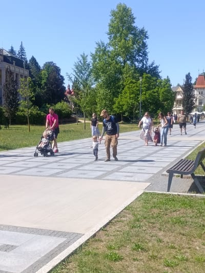 The image depicts a sunny day scene in Luhačovice, Czechia, specifically along a paved walkway in what appears to be a park or promenade. A man in a bright pink shirt is pushing a stroller with a young child in it. Another young child is walking alongside him. Further down the path, several other individuals, including adults and children, are seen walking in the same direction. The environment includes manicured green lawns, trees, and buildings in the background, suggesting a well-maintained public space. The architecture of the buildings hints at a European setting, consistent with the reported location.