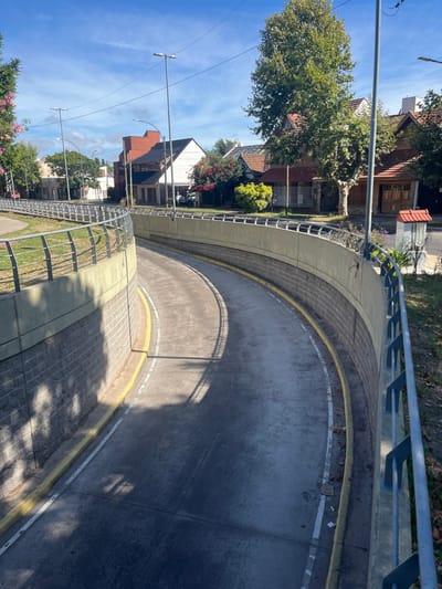 A curved road runs between a sidewalk and a retaining wall with a metal railing. Sunlight creates shadows across the gray road. Houses and trees are visible beyond the wall and railings, set against a blue sky with a few clouds. Streetlights line the sidewalk. The road appears to be heading underground.