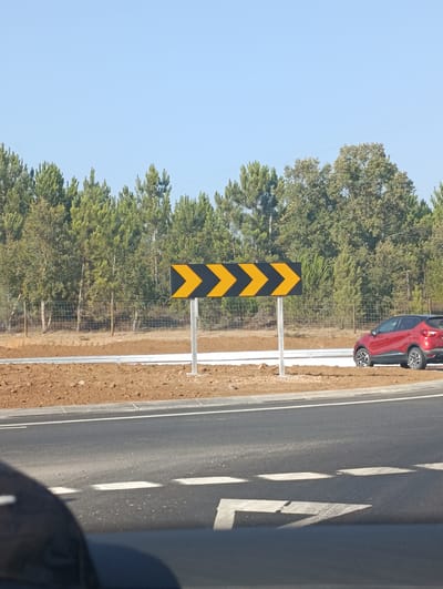 The image shows a road sign with multiple yellow arrows pointing to the right, indicating a turn. The sign is black and yellow and is mounted on a metal pole. A red car is parked on the right side of the road. The background consists of trees and a clear blue sky. The scene is likely located in the city of Santiago do Cacém, Portugal, as suggested by the context.