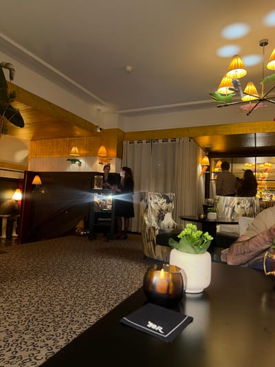The image shows the interior of a dining establishment, possibly a restaurant or upscale bar in Bellevue, Switzerland. The camera angle is skewed, with a focus on the right side of the frame.
**Environment:** The interior features a mix of modern and classic decor. The ceiling is predominantly white, with some sections of gold-colored paneling. A chandelier with multiple arms and unique light fixtures hangs from the ceiling. The floor is covered in a leopard-print carpet.
**Objects:** Visible objects include tables, chairs with patterned upholstery, decorative plants, a candle holder, a menu or bill holder, and a bar with bottles displayed in the background.
**People:** Two women are standing near a service station and looking at something with bright light. There are other patrons and staff members visible in the background.
**Actions/Interactions:** The people in the background appear to be seated and engaged in conversations or dining. The two women in the foreground appear to be reviewing something that casts a bright light.
**City of Bellevue, Switzerland:** Given the upscale decor and ambiance, this establishment is consistent with the type of businesses found in Bellevue.