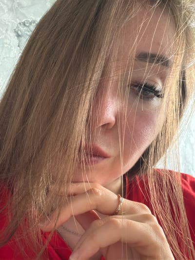 A close-up, waist-up shot shows a young woman with long, light brown hair that partially obscures her face. Her head is tilted, and her chin rests on her hand, which is adorned with a ring. She is wearing a red top. The background appears to be a textured wall, possibly with a lace pattern. The lighting suggests an indoor setting. There is no information to determine the location as Старые Туймазы, Russia.