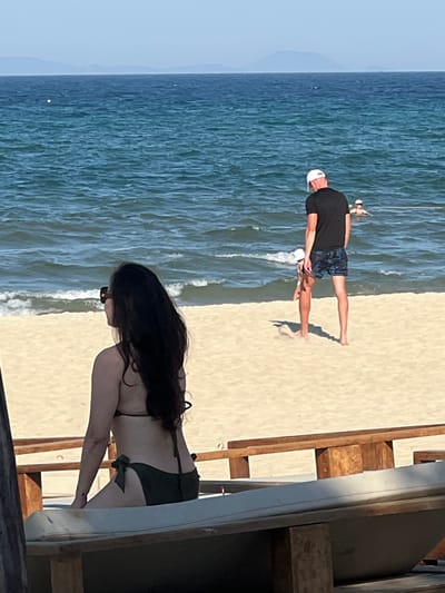 This image depicts a beach scene in Đà Nẵng, Vietnam. In the foreground, a woman with long dark hair is seated, facing away from the camera, wearing a dark green bikini. She is positioned on a lounge chair, with a wooden railing in front of her. In the middle ground, on the sand and at the edge of the water, a man wearing a black t-shirt and patterned swim shorts stands. A young child, wearing a white hat, is near the man, partially obscured. Further out in the blue ocean, another person is visible swimming. The background shows a hazy horizon with distant landforms. The sand is light brown, and the water is a deep blue with small waves.