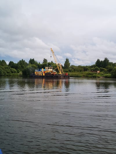 The image shows a crane barge on a body of water, likely a river, with a densely wooded shoreline in the background. The barge is yellow and black, with a tall yellow crane mounted on it. The barge appears to be stationary. The water is rippled, suggesting some movement. The sky is overcast with clouds. The general impression is one of an industrial or work barge on a natural waterway. The location is identified as Kargulino, Russia.