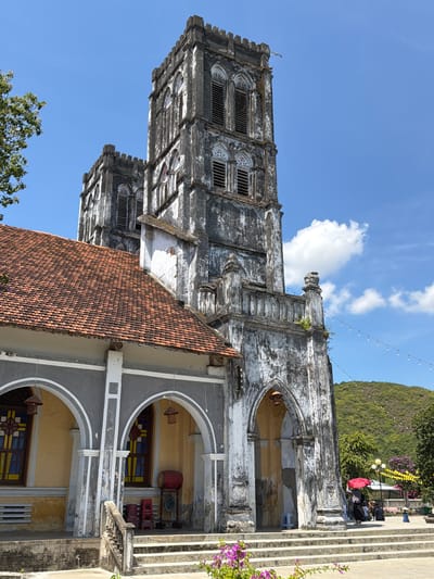 The image depicts the exterior of a weathered, old church with two prominent towers. The architecture suggests a Gothic or Neo-Gothic style, characterized by arched windows, decorative stonework, and crenellations on the towers. The building's facade shows signs of age, with peeling paint and discoloration, indicating it has stood for a considerable time.

The left side of the building features a tiled roof with orange terracotta tiles. Beneath the roof, a section of the building is painted gray with white trim, housing arched doorways or windows with decorative stained glass. In front of these arches, there are several red plastic chairs and a large red drum, possibly a musical instrument or decorative element.

The central part of the image is dominated by the church's towers. The taller tower on the right has multiple levels of arched openings, some of which are filled with louvers, suggesting they are bell towers. The stonework is heavily textured and stained, with patches of white paint contrasting with darker, aged stone. A small balcony or walkway with a balustrade runs across the mid-section of the tower.

The ground in front of the church is paved with concrete and features a set of wide, stone steps leading up to the entrance. A stone railing lines the steps. In the foreground, on the steps and to the left, are clusters of vibrant purple flowers.

In the background, lush green hills rise behind the church. The sky is a clear, bright blue with a few fluffy white clouds. String lights are visible hanging across the scene, adding a festive or decorative element. Several people are present near the base of the church, some seated, and one individual is holding a bright red umbrella. The overall impression is of a historical landmark under a bright, sunny sky. The location is identified as Hội Tín, Vietnam.
