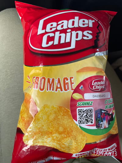 The image shows a close-up view of a sealed red and yellow bag of "Leader Chips" potato crisps. The red top section displays "Leader Chips" in white, while the central yellow section features "FROMAGE" (Cheese) in orange text. Golden-brown potato chips are illustrated across the lower yellow portion of the bag. A circular red sticker on the right side contains a "Leader Chips" logo, a text field with the code "DAQ3E4KD," a QR code labeled "SCANNEZ" (Scan), and the phrase "ET TENTEZ DE GAGNER" (And try to win), accompanied by illustrations of a gaming PC tower, a laptop, a smartwatch, and a smartphone as prizes. Partially visible text at the bottom states "POMMES DE TERRE DU MAROC" (Potatoes from Morocco) and "SANS CONSERVATEURS" (Without preservatives). The background consists of a blurred, light-colored fabric, consistent with upholstery or clothing. The scene is located in Sidi El Makhfi, Morocco.