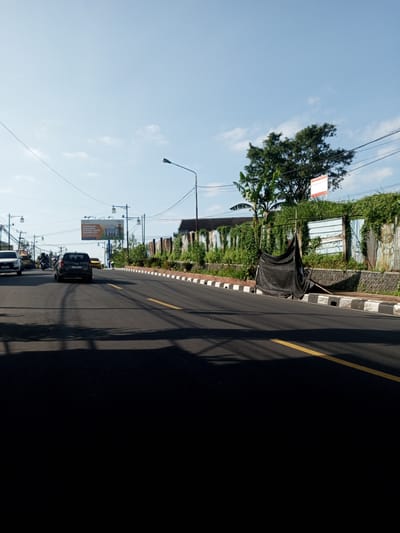 The image captures a street scene in daylight. The foreground is mostly dark, presumably under a bridge or overhang, obscuring details. A two-lane road with yellow lines runs through the center of the frame, with vehicles traveling on the left side. A car is visible heading away from the camera. The road is lined with a white and black curb on the right side. A weathered fence, overgrown with greenery, runs parallel to the road on the right. A black tarp hangs from a makeshift structure next to the fence. Streetlights are visible along the road, as is a billboard in the distance. The sky is blue, with a few clouds. There are no indications of it being Nhot Chang, Laos.