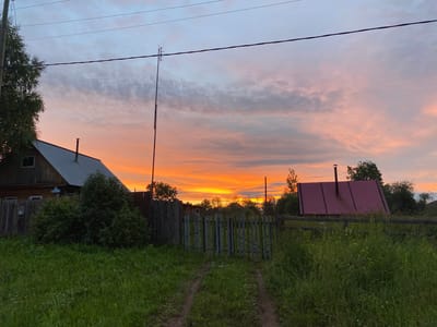 The image depicts a rural scene in Баклуши, Russia, at dusk. The sky is filled with vibrant hues of orange, pink, and purple, indicating a beautiful sunset. Wispy clouds are scattered across the sky, catching the last rays of sunlight.
In the foreground, a grassy field with a dirt path leading through it dominates the lower portion of the frame. The path shows tire tracks, suggesting recent vehicle use. A rustic wooden fence runs horizontally across the middle ground, with a gate in the center.
To the left, a wooden house with a metal roof is visible, partially obscured by lush green bushes and trees. A chimney protrudes from the roof. To the right, another structure with a distinctive red metal roof is seen, also surrounded by greenery. A tall pole with an antenna stands near the center of the image, with power lines stretching across the sky.
The overall mood is peaceful and serene, evoking the quiet beauty of a rural evening. The time of day is clearly sunset, and the weather appears calm, given the soft lighting and clear sky. There is no visible text in the image.