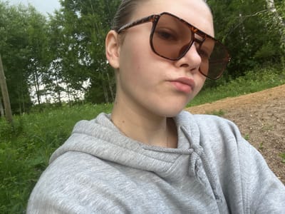 A young person with blonde hair pulled back is shown in a close-up, selfie-style photograph. They are wearing large, tortoiseshell-patterned sunglasses that cover a significant portion of their face, and a light gray hooded sweatshirt. The person is looking directly at the camera with a slightly pursed-lip expression. A delicate silver necklace is visible around their neck.
The background suggests an outdoor setting. Lush green trees and foliage form a dense backdrop, with some lighter patches indicating the sky or a clearing. The ground in the foreground appears to be a mix of dirt and dry leaves. The overall lighting suggests it is daytime, possibly late afternoon or early evening, with soft light and overcast skies. The environment gives the impression of a park, woods, or a rural area. There is no visible text in the image. The subject's pose and direct gaze indicate a casual, personal moment captured by the camera.