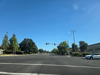 This media file captures a street scene in Elk Grove, USA, on a clear, sunny day, likely mid-morning or early afternoon given the bright sunlight and long shadows. The perspective is from a vehicle traveling on a paved road, looking forward at an intersection.
The primary subjects are the road, traffic signals, street signs, trees, and a white electric car. The road has clear lane markings and is in good condition. Several traffic signals are visible, with one showing a red light and a left-turn arrow, indicating a stop or a turn is being controlled. Another signal to the right also shows a red light. A street sign displays "Poppy Ridge" and "Quail Run," providing location context.
Lush green trees line the sides of the road, contributing to a suburban or park-like atmosphere. On the right, a modern building with a light-colored facade is visible. A white electric car, possibly a Tesla, is turning right at the intersection.
The scene appears calm and ordinary, with no people prominently visible. The weather is fair and the sky is a vibrant, cloudless blue. The overall impression is one of a typical day in a well-maintained suburban environment. The activity is the controlled movement of traffic through an intersection.