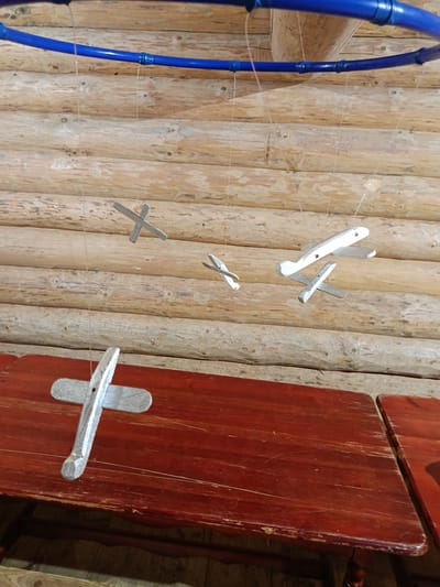 In Semenkovo, Russia, within a log cabin structure, a mobile consisting of several small, silver-painted wooden airplane models hangs from thin, transparent threads. These threads are attached to a blue, plastic hoop that is suspended from the ceiling. The log walls of the cabin are visible in the background. In the foreground, a dark red wooden table with visible scratches and wear is positioned beneath the mobile.