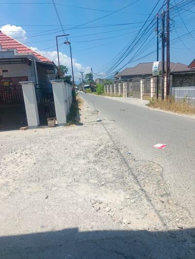 The image depicts a street scene in Tenggela, Indonesia, under a clear blue sky with scattered clouds. The foreground shows a rough, gravelly road surface. To the left, a house with a red tiled roof and a white fence is visible. A lamppost stands near the house. Further down the road, on the right side, a fence with decorative metalwork borders a property with a house. A power pole with numerous wires stretches across the scene. The road continues into the distance, where a few vehicles and buildings can be seen. A red and white object, possibly a piece of paper or a flyer, lies on the road surface. The overall impression is of a typical residential street in a sunny, tropical environment.