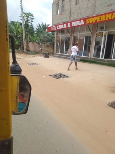 The image depicts a street scene in Achalla, Nigeria. The view is from a vehicle, with a yellow frame and a rearview mirror visible on the left foreground; the mirror reflects an occupant and interior elements. A two-story building anchors the right side of the frame. Its upper floor is unfinished, exposing concrete block construction and open window spaces. The ground floor houses "ZARA & MIRA SUPERMARKET," indicated by a prominent red sign with white lettering across a facade composed of multiple glass storefront panels and doors.
A sandy, unpaved road occupies the center and middle ground, with a narrow strip of paved road visible on the far left. Two square metal drainage grates are embedded in the sandy road surface. On the dirt verge outside the supermarket, near the left side of its entrance, a yellow and black portable generator is visible. Further to the right, a roll of green artificial turf leans against the lower part of the supermarket's facade.
A single individual, wearing a white t-shirt and patterned shorts, walks barefoot or in sandals across the sandy road, moving away from the camera towards the supermarket entrance. In the left background, dense tropical vegetation, including banana trees and palm trees, grows behind a low wall. Utility poles and power lines are visible against an overcast sky.