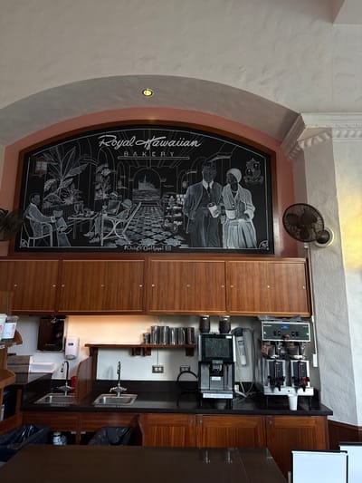 The image shows the interior of the Royal Hawaiian Bakery in Honolulu, USA. A large, arched chalkboard to the left depicts a scene of the Hawaiian monarchy, the islands, and tropical flora. A wooden cabinet and countertops on the right hold coffee-making equipment, sinks, and other bakery supplies. A ceiling fan is mounted near the top center of the image.