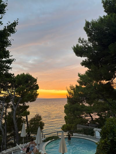 The image depicts a sunset over the Adriatic Sea, viewed from a terrace in Općina Brela, Croatia. The sky is a gradient of pale blue and soft orange hues, with wispy clouds. Two people are visible near a circular swimming pool filled with clear blue water. One person, wearing a floral top and denim shorts, is sitting on a lounge chair, looking down. Another person, with long dark hair, is partially submerged in the pool, with their back to the camera, appearing to wave an arm. Two large, cream-colored beach umbrellas stand near the lounge chairs. Lush green pine trees frame the scene, with their branches reaching towards the sky. The sea stretches out to the horizon, reflecting the warm colors of the sunset. A stone wall and some greenery form the edge of the terrace.