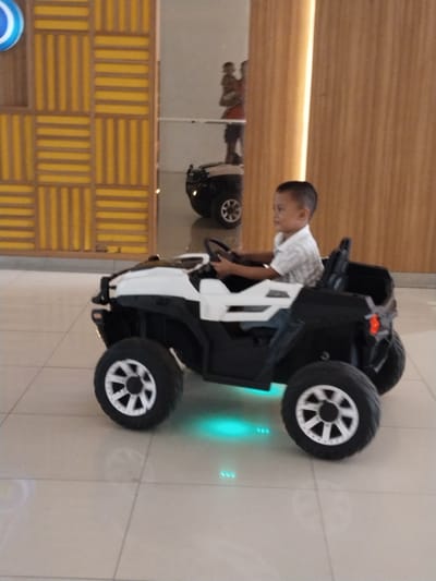 A young boy of Asian descent, wearing a light-colored, short-sleeved collared shirt and dark trousers, is seated in the driver's position of a black and white toy ATV. He is holding the steering wheel, appearing to be in motion. The ATV has large black tires with silver-rimmed wheels and features illuminated blue lights underneath. The background includes a patterned wall with yellow and wood tones and a blurred reflection of other people and a similar toy vehicle. The scene appears to be set indoors, possibly in a shopping mall or entertainment venue in Kudus, Indonesia.