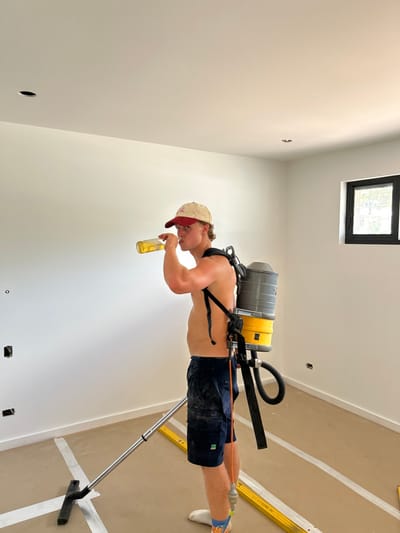 A shirtless man wearing a baseball cap and a backpack vacuum cleaner drinks from a beer bottle. He stands in a room with white walls and a light brown floor marked with white lines. A window is in the upper part of the frame. A long tool with a flat head rests on the floor, and a level is visible on the right. The image has been rotated 90 degrees counter-clockwise.