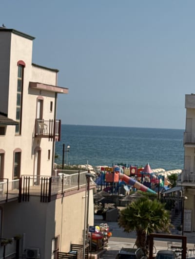 This is a medium-distance, eye-level shot of a coastal scene in Rimini, Italy. The foreground is dominated by the side of a cream-colored, multi-story building with brown trim and balconies. A second building, partially visible on the right, is also light-colored with balconies. Beyond the buildings, the blue sea stretches to the horizon, meeting a clear sky.
In the midground, a vibrant children's playground with colorful structures, including slides and climbing frames, is visible near the beach. Several beach umbrellas are scattered on the sandy area. A palm tree stands in front of some buildings on the right side of the image. A street lamp is visible between the buildings and the playground.
The image appears to have been taken on a sunny day, likely during the summer season, given the beach and playground setting. There are no people prominently featured in the image. The overall atmosphere is one of a sunny, tourist-oriented seaside location.