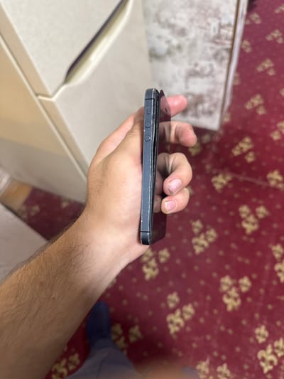 A person's hand is holding a dark-colored smartphone, viewed from the side, in what appears to be an indoor setting. The phone's side buttons and frame are visible. The background consists of a beige cabinet or appliance and a red carpet with a floral pattern. The scene suggests a casual or domestic environment.