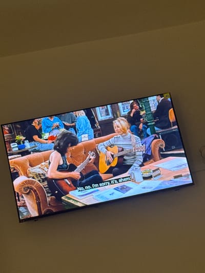 The image shows a television screen displaying a scene from the TV show "Friends." Two women are seated on a couch, each playing a guitar. The woman on the left has dark hair and is wearing a black tank top. The woman on the right has blonde hair and is wearing a striped sweater. In the background, other people are visible sitting on a couch and at a table. The room appears to be a cafe or apartment setting. There is text on the screen that reads "No, no. I'm sorry, it's, ahem:". The photograph is taken from an angle, suggesting it is a candid shot of the TV screen. There is no indication or information within the image that connects it to the city of Naousa, Greece.