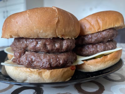 The image shows two double cheeseburgers on a black plate. The burgers are stacked high with multiple patties and slices of cheese. The buns are golden brown and appear soft. The burgers are on a table covered with a patterned cloth. The background is blurred and appears to be an indoor setting, possibly a kitchen or dining area. There are no people or animals visible in the image. There is no information about the location in the city of Annalong, United Kingdom.