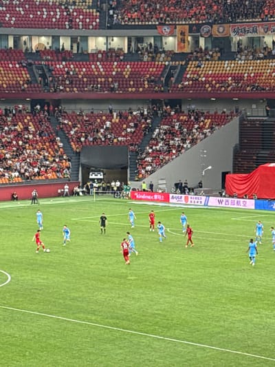 The image shows a soccer game at the Chongqing Longxing Football Stadium in Longxing, China. The stadium is full of fans seated in red and yellow seats on one side. On the field, players in red and blue uniforms are actively playing. A large red banner is displayed next to the field. There are also advertising billboards along the side of the field. There are referees on the field as well.