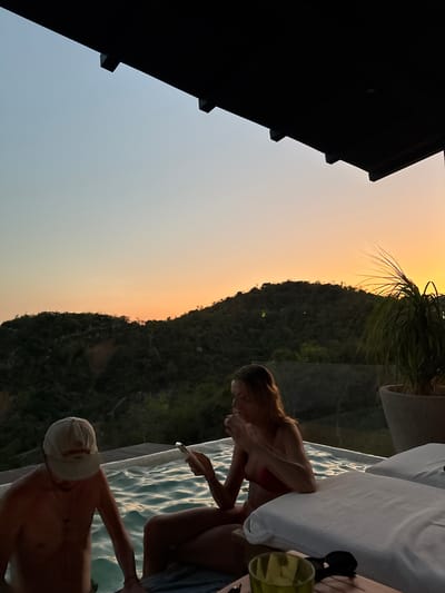 The image shows a sunset scene at a pool, likely in a tropical or coastal location.
People: A woman in a red swimsuit is visible, holding a smartphone. A man wearing a white cap is in the pool.
Objects: Pool with light blue water, white lounge cushion, green plant, clear glass.
Environment: Green mountains are in the background, and the sky has hues of orange and blue, indicating sunset. The location may be in Puerto Marqués, Mexico, given the foliage and the general landscape. The edge of a dark roof is visible on the left side of the image.