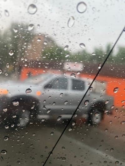 This image captures a scene viewed through a rain-streaked window, likely from inside a vehicle. The foreground is dominated by numerous water droplets on the glass, distorting the view beyond. In the mid-ground, a silver pickup truck is visible, with its headlights illuminated. Behind the truck, a building with an orange facade and some trees can be seen. The overall atmosphere is overcast and appears to be during daylight hours. The location is specified as Hromtau, Kazakhstan, implying this is a street scene within that city. There are no people clearly visible in the image.