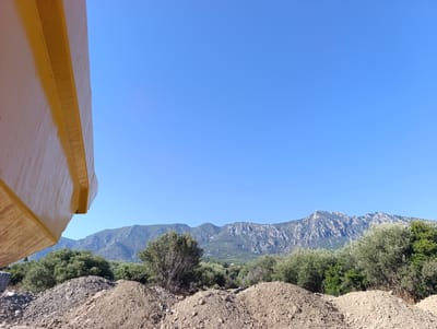 A bright, clear blue sky dominates the upper portion of the image. In the foreground, a large, textured pile of dirt and rocks is visible. To the left, a portion of a yellow, angled object, possibly part of construction equipment, is in sharp focus. Behind the dirt pile, a dense line of green trees and bushes stretches across the mid-ground. Further in the distance, a rugged mountain range with rocky, uneven peaks and slopes is silhouetted against the sky. Some lighter green patches, suggesting vegetation or buildings, are scattered on the lower slopes of the mountains. The overall scene appears to be an outdoor location, likely a construction site or undeveloped area, with a mountainous backdrop. The location is stated to be Erdemit, Northern Cyprus / Cyprus. There are no people or animals visible in the image.