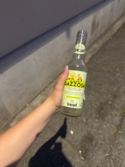 A hand is holding a bottle of Lurisia Gazzosa lemon soda. The bottle is mostly empty. The background shows an asphalt road or sidewalk next to a concrete wall.