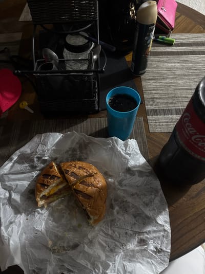 A table arrangement includes a cut hamburger wrapped in white paper, a blue cup filled with dark liquid, and a bottle of Coca-Cola. Beside these are a pink wallet, a can, and a black metal basket containing utensils. A grey placemat sits beneath some of the items. A red cap is near a yellow key at the bottom corner.