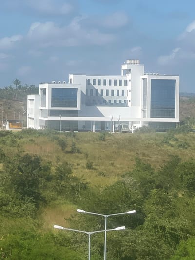 The image depicts a modern, multi-story white building with numerous windows and glass facades, set against a backdrop of lush green trees and a pale blue sky with scattered clouds. The building's architecture suggests it is an office building or a hotel, with sharp, clean lines and a contemporary design. There are no people visible in the image, but the presence of streetlights on poles in the foreground indicates it is an outdoor scene. The lighting suggests it is daytime, likely morning or afternoon, with the sun casting some shadows on the building. The overall setting appears to be a developed area surrounded by natural vegetation, possibly in a suburban or semi-rural location. No text is visible in the image. The scene conveys a sense of order and modernity within a natural environment.