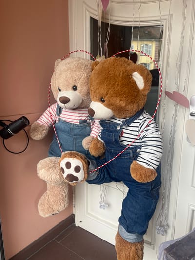 The scene displays two large plush teddy bears positioned upright, facing slightly forward, against a light peach-colored wall on the left and a white, paneled exterior door on the right, in the city of Sélestat, France. The door's glass reflects a light-colored building with dark roofing and windows across a street.

The left bear is tan-colored, wearing a red and white striped long-sleeve shirt under blue denim overalls. Its right paw is visible, showing a brown pad with white outlines. The right bear is dark brown, dressed in a black and white striped long-sleeve shirt and blue denim overalls. A red and white striped cord forms a heart shape around the heads of both bears.

To the upper-left of the tan bear, a black cylindrical object, resembling a small speaker or camera, is mounted on the peach wall, connected by a black cord. The white door is adorned with multiple transparent strings holding small, clear decorative lights and several pink and white paper heart cutouts suspended by strings. A dark brown baseboard runs along the bottom of the peach wall. The ground is a dark, tiled surface.