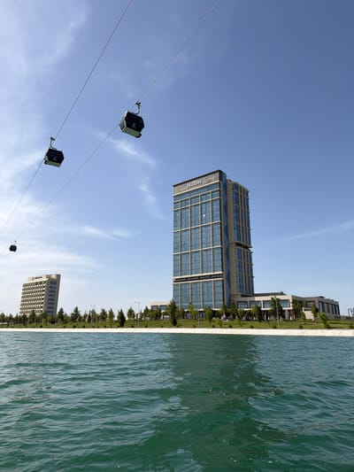 The image displays a modern urban landscape under a bright, clear blue sky. A prominent, multi-story building featuring a glass and metal facade is visible, with "Hilton" clearly legible on its side, indicating it is a hotel. Other contemporary buildings are situated further along the waterfront, some with similar architectural styles.
In the foreground and extending into the midground, a wide body of turquoise-green water, resembling an artificial lake or canal, stretches across the scene. Its surface shows subtle ripples from gentle movement. A clean, light-colored concrete edge borders the water, separating it from a line of small green trees and shrubs planted along the bank.
Above, against the clear sky, a cable car system is visible, with three dark-colored cabins suspended from taut wires that span diagonally across the frame. The sky is a vibrant blue with only a few faint, wispy clouds. The strong, even lighting suggests it is midday or early afternoon on a sunny day. No people are visible in the scene. The overall impression is one of a clean, modern, and developed leisure or urban area. The location context is Kuchtumgali, Uzbekistan.