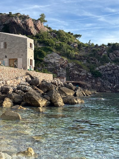 This is a medium-angle, natural light, eye-level shot of a coastal scene in Rijeka Reževići, Montenegro. On the left, a light-colored stone building with white shutters on its windows sits on a rocky slope. A low stone wall, constructed with small, irregularly shaped stones, separates the building from a jumble of large, grey rocks that spill down to the water's edge. The water is clear, revealing a pebbled seabed and reflecting the sunlight. Behind the building, the rocky hillside is covered in dense green vegetation. In the background, a larger, rugged mountain range with exposed pink and grey rock formations rises against a blue sky dotted with thin, white clouds. To the right of the building, further down the coastline, a small, rustic structure with a thatched roof and a fishing net draped over it is nestled amongst the rocks. The scene depicts a quiet, natural coastal environment with a touch of human habitation.
