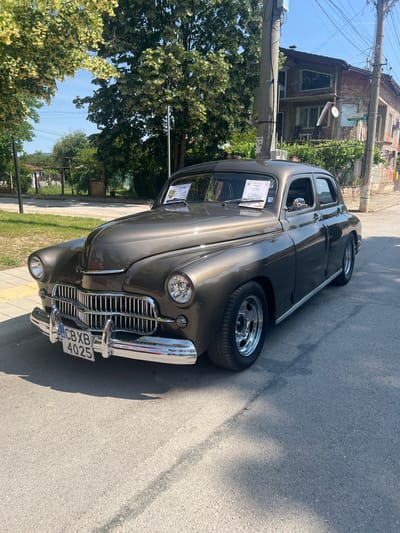 A vintage car, a metallic brown sedan, is parked on an asphalt road in front of a two-story brick building. The car, featuring chrome accents and wire-spoke wheels, has its license plate displayed, reading "BG CXB 4025". A sign on the car's windshield indicates it is participating in an event. The background includes a tree with yellow blossoms, more trees, power lines, and a partially visible building with a Bulgarian flag hanging from its facade. The location is identified as Skravena, Bulgaria.