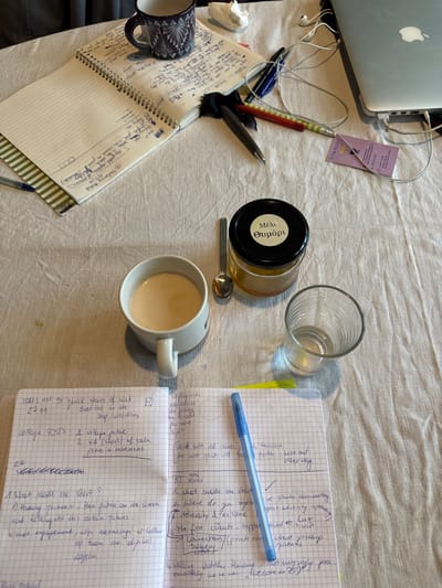 The image displays an overhead view of a cluttered work surface on a light-colored, possibly linen, tablecloth, located in Saint-Gilles - Sint-Gillis, Belgium.
In the upper-left quadrant, a dark blue ceramic mug with a geometric pattern is situated next to an open spiral-bound notebook containing handwritten notes in blue ink. A crumpled white tissue, three pens (one black, one red-and-white striped, one dark blue), and an earbud cable are scattered nearby.
To the upper-right, a silver laptop, likely a MacBook, is partially visible, connected to white wired earbuds and other cables. A small purple business card with text in a light color rests beneath the earbud cable.
Centrally positioned, a white ceramic mug contains a light brown liquid, possibly tea or coffee with milk. A small silver spoon rests beside it. To the right, a clear glass jar with a black lid is present, labeled "Μέλι Θυμάρι" (Thyme Honey), filled with a golden, viscous substance. An empty clear glass with vertical ridges stands adjacent to the honey jar.
In the foreground, a second open spiral-bound notebook with grid paper is filled with handwritten notes in blue ink. Visible text includes "COLLAGE POSTS:" and "What made me start?". A blue ballpoint pen lies diagonally across the right page of this notebook, with a small yellow sticky note partially visible beneath it. The items are static, suggesting a paused work or study session.
