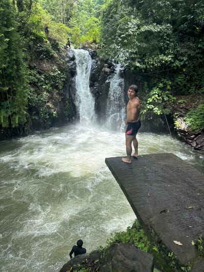 The image depicts a natural waterfall scene in Ambengan, Indonesia, characterized by dense, lush green tropical foliage. In the foreground, a shirtless male with dark hair, wearing black swim trunks with red and white accents, stands barefoot on a grey concrete diving platform that extends over a turbulent body of water. He is positioned with his back partially to the camera, looking over his right shoulder towards the viewer. A prominent waterfall cascades from a rocky cliff face in the midground, creating a foamy, light-green pool of water below. To the lower left, another individual in a dark long-sleeved shirt is partially visible, standing on a dark rock at the water's edge. In the upper left background, two additional individuals are observable on the rocky terrain above the waterfall. The environment is rich with varied vegetation, including large trees, smaller plants, and bamboo stalks.