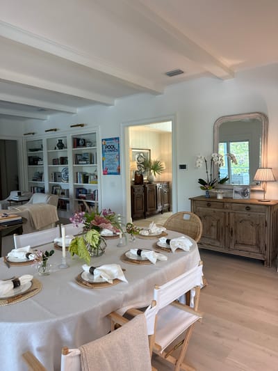 The image depicts an interior scene of a dining room, likely in East Naples, Florida, given the style of the architecture and decor. A large, oval dining table is centrally positioned, covered with a light-colored linen tablecloth. The table is set for a meal with placemats, plates, white napkins with dark napkin rings, and cutlery. A floral arrangement in a glass vase and several candles adorn the table. White director's chairs with light-colored canvas seats are placed around the table.
In the background, a white wall features a large built-in bookshelf filled with books and decorative items. To the right of the bookshelf, a framed "POOL OLYMPICS" poster is displayed. Further to the right, a distressed wooden sideboard stands against the wall, topped with a large ornate mirror. On the sideboard, there are decorative objects including a lamp with a pleated shade, a vase with orchids, a white bowl, and a framed photograph. Through an open doorway to the left, another room with light-paneled walls and a wooden cabinet is visible. The room has a light-colored wood floor and a coffered ceiling with white beams. No people are present in the image.