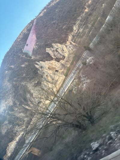 The image depicts an oblique, elevated view of a rugged landscape in the vicinity of Struya, Bulgaria, under clear daylight conditions. A steep hillside, covered predominantly with bare deciduous trees and sparse brown vegetation, occupies the majority of the frame. In the mid-ground of the hillside, a series of light-colored, weathered rock formations with jagged contours and several dark, rectangular openings, possibly cave entrances or shelters, are prominent. Higher on the slope, partially integrated into the terrain, stands a distinctive, pale pink-colored structure featuring a single dark rectangular opening. Below the rocky formations, a multi-lane paved road with visible lane markings and guardrails traverses horizontally across the scene. Parallel to and adjacent to this road, a set of parallel tracks, consistent with a railway line, is present. Additional infrastructure, appearing to be a segment of a bridge or an underpass, is partially visible at the bottom of the frame. The sky in the upper left quadrant is clear blue. No individuals, vehicles, or active discernible events are present.