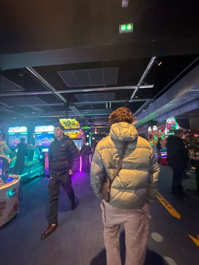 The image depicts an indoor entertainment venue, specifically an arcade, identified as Bowling Saint André - Laser Game & Arcades, located at Zone Industrielle de la Chapelle, Rue du Bosphore, 59350 Saint-André-lez-Lille, France.
The scene is illuminated by the vibrant, multi-colored lights emanating from numerous arcade machines lining a central walkway. The overall lighting is dim, contrasting with the bright game displays. The ceiling is dark, featuring visible grid panels, ventilation ducts, and a green illuminated exit sign showing two running figures positioned in the upper right. The floor is covered with a dark blue or grey carpet, marked with a yellow curved arrow and the partial French text "C'est déjà ça" on the right side.
In the foreground, a male subject is shown from behind, walking away from the camera. He has curly brown hair and wears a light olive-green puffer jacket, light-colored cargo pants, and a dark brown leather messenger bag slung across his shoulder. In the midground, slightly left of center, another male subject is walking towards the camera. He has short, dark hair and wears a black puffer jacket with an Under Armour logo on the left chest, dark pants, and dark sneakers with reddish soles; his right hand is slightly clenched.
Multiple other individuals are visible in the background; some are indistinct figures near or interacting with arcade machines. Visible game titles on the machines include "WAVE RIDERS" and "SUPER KICKS 3". No direct interaction between the primary subjects is occurring; both are in motion.