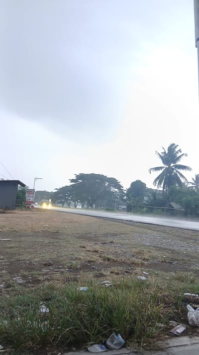 This media file depicts an outdoor scene in Sumbokrayek, Indonesia. The image is oriented vertically, with the sky dominating the left side and a roadside area on the right.

The sky is overcast and appears to be a bright, hazy white, suggesting either early morning, late afternoon, or a very cloudy day. The weather seems to be rainy, as indicated by the wet appearance of the road and the general atmospheric conditions.

On the right side of the image, a paved road runs diagonally. Adjacent to the road is a grassy area with patches of dry earth and scattered stones. Several pieces of litter are visible in the grass and along the roadside, including plastic bags and bottles.

Further back, along the road, are several trees, including a prominent palm tree with its fronds spread out, and a large, leafy tree. In the distance, two yellow headlights are visible on what appears to be a vehicle on the road. A small building with a dark roof is also visible in the lower right portion of the frame, and a sign attached to a pole nearby displays some text. Partially visible text on the sign includes "Canon," "Cuts Care," and a phone number.

The overall mood of the image is somewhat somber due to the weather and the presence of litter, but the bright headlights in the distance offer a hint of activity and movement within the scene. The location cues, such as the palm tree and the type of vegetation, are consistent with a tropical or subtropical setting like Indonesia.