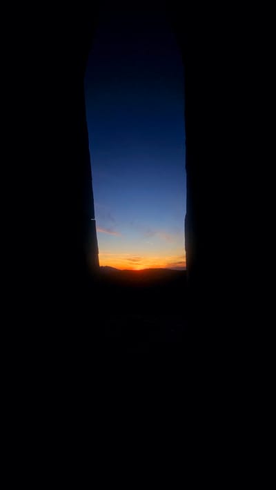 A dramatic sunset is framed by the dark silhouettes of two vertical structures, possibly ancient stones or architectural elements, that create a narrow, arch-like opening. The sky transitions from a deep indigo at the top to vibrant orange and yellow hues near the horizon, where a silhouetted mountain range is visible. Thin, wispy clouds catch the last rays of sunlight. The foreground and immediate surroundings within the opening are obscured by shadow, suggesting the image was taken from a dark interior looking out. The overall mood is serene and awe-inspiring, with the natural beauty of the sunset contrasting with the stark, man-made or natural framing. The location is identified as Simeonovets, Bulgaria.