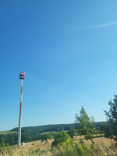 This media file captures a clear, bright day with a vast expanse of blue sky dominating the frame. In the foreground, a tall, slender tower with red and white banding extends diagonally upwards. At its apex, a complex structure with multiple levels and visible metal components suggests it's a telecommunications tower or similar infrastructure.

The background reveals a pastoral landscape, indicative of a rural setting. A sloping field, likely agricultural land, is dotted with what appear to be round hay bales. Beyond this, a dense line of green trees forms a natural barrier, suggesting a forest or wooded area. The edge of the frame on the right side shows more vegetation, possibly grasses or hedges, and hints at a roadside or field boundary.

The lighting and the clear blue sky indicate a time of day likely late morning or early afternoon, with sunny weather. The scene evokes a sense of openness and tranquility, common to rural German landscapes. There is no visible text in the image. The orientation of the image is such that the tower appears to be in the lower right quadrant, and the landscape is visible in the upper right quadrant.