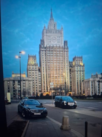 The image depicts a twilight scene in a city, featuring a prominent Stalinist skyscraper in the background. The skyscraper, likely a government building due to its style and imposing architecture, is illuminated, with visible windows and decorative elements. In the foreground, a black Mercedes-Benz sedan is parked on the right side of the street, facing forward. To the right of the sedan, a black van is driving on the road. A street lamp with two lights is visible to the left of the skyscraper, casting an orange glow. The sky is a deep blue, suggesting either dawn or dusk, with some faint clouds. The area appears to be a public space with a sidewalk and a street. There is no indication that this scene is located in Užuežerė, Lithuania; the architecture is characteristic of Moscow or other cities in former Soviet republics.