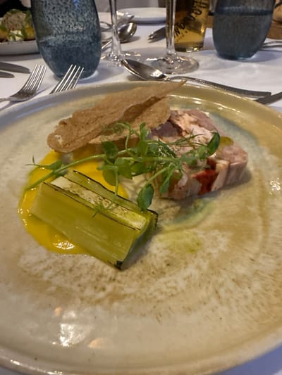 The image shows a plate of food on a table setting. The plate contains sliced meat, a charred leek, fried bread, yellow sauce, and a green garnish. Cutlery and glassware are visible on the table. The location is not specified in the image, so it cannot be confirmed to be in Glenridding, United Kingdom.
