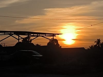 A sunset is depicted in Cot Buket, Indonesia. The sky is a gradient of yellow and orange, with thin clouds stretched across. Silhouetted in the foreground are trees and buildings, an open-roofed structure on stilts, and a car. Power lines extend across the upper portion of the frame, with a bird perched on one.