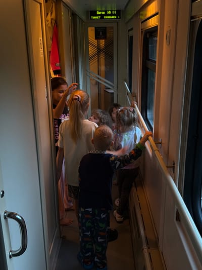A group of children and adults are in a train compartment. A digital sign above a doorway indicates "VAGON NO. 11", with Russian text below it that appears to mean "FREE". The compartment is narrow with a window on the right side. Several people, including women and children, are visible, some looking out the window and others interacting with each other. One child is wearing pajamas with a dinosaur print. A metal handrail runs along the window. There is a closed door on the left with a large silver handle. In the background, behind a doorway, a red jacket and a white shirt are hanging. The overall lighting suggests it is either early morning or evening, with a warm glow from the train's interior lighting. The specific location of Troekurovo, Russia, is not depicted in the visual elements of the image.